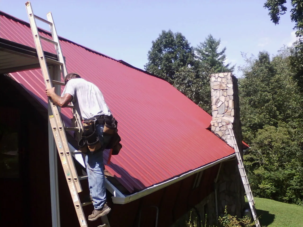 Roofer on a ladder beside a red metal roof and stone chimney — Soffit & Fascia Repair by Roofing Brothers in Orovada